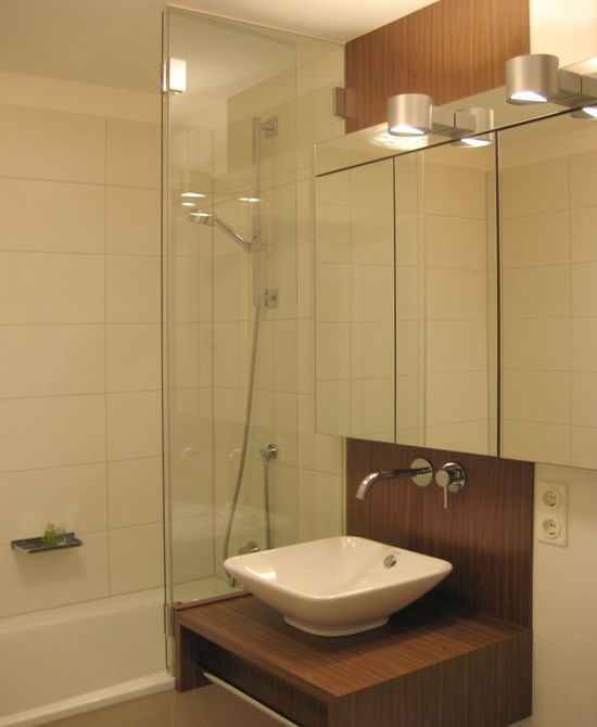 Der freihängende Waschtisch mit Aufsatzbecken, Wandarmatur und Spiegelschrank schließt an die Duschabtrennung aus Glas an. Diese steht auf dem Rand der Badewanne auf..