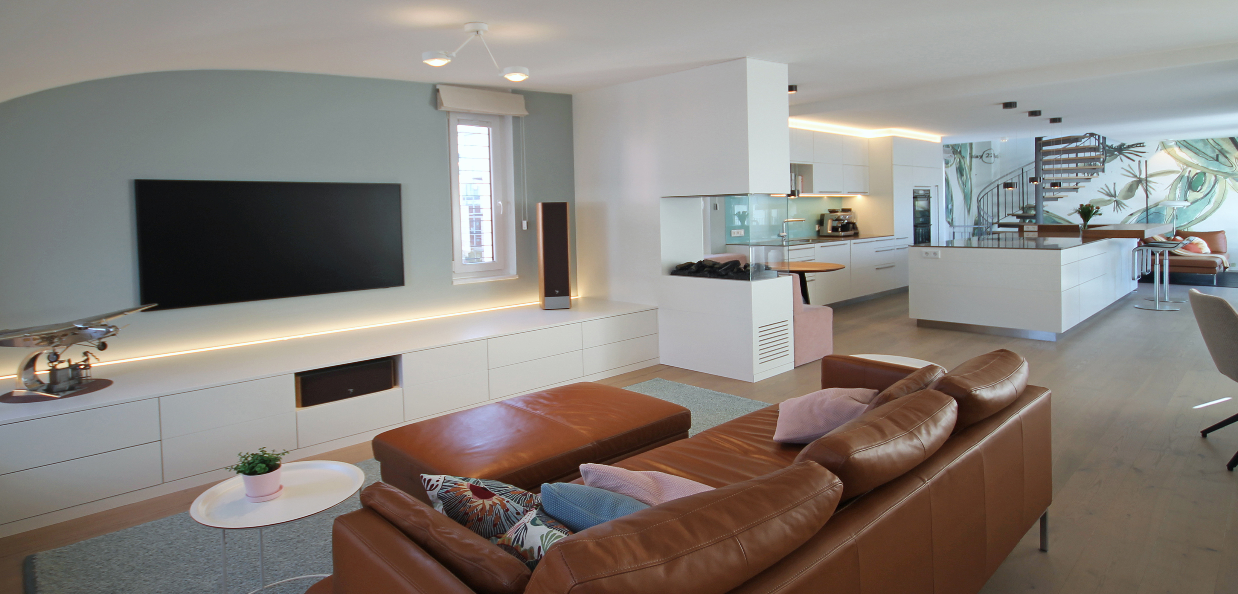 Modern gestalteter Wohn‑ und Küchenbereich einer Maisonettewohnung mit hellen Wänden, eingebauter Medienwand, braunem Ledersofa, minimalistischer Einbauküche und offener Treppe ins Obergeschoss.