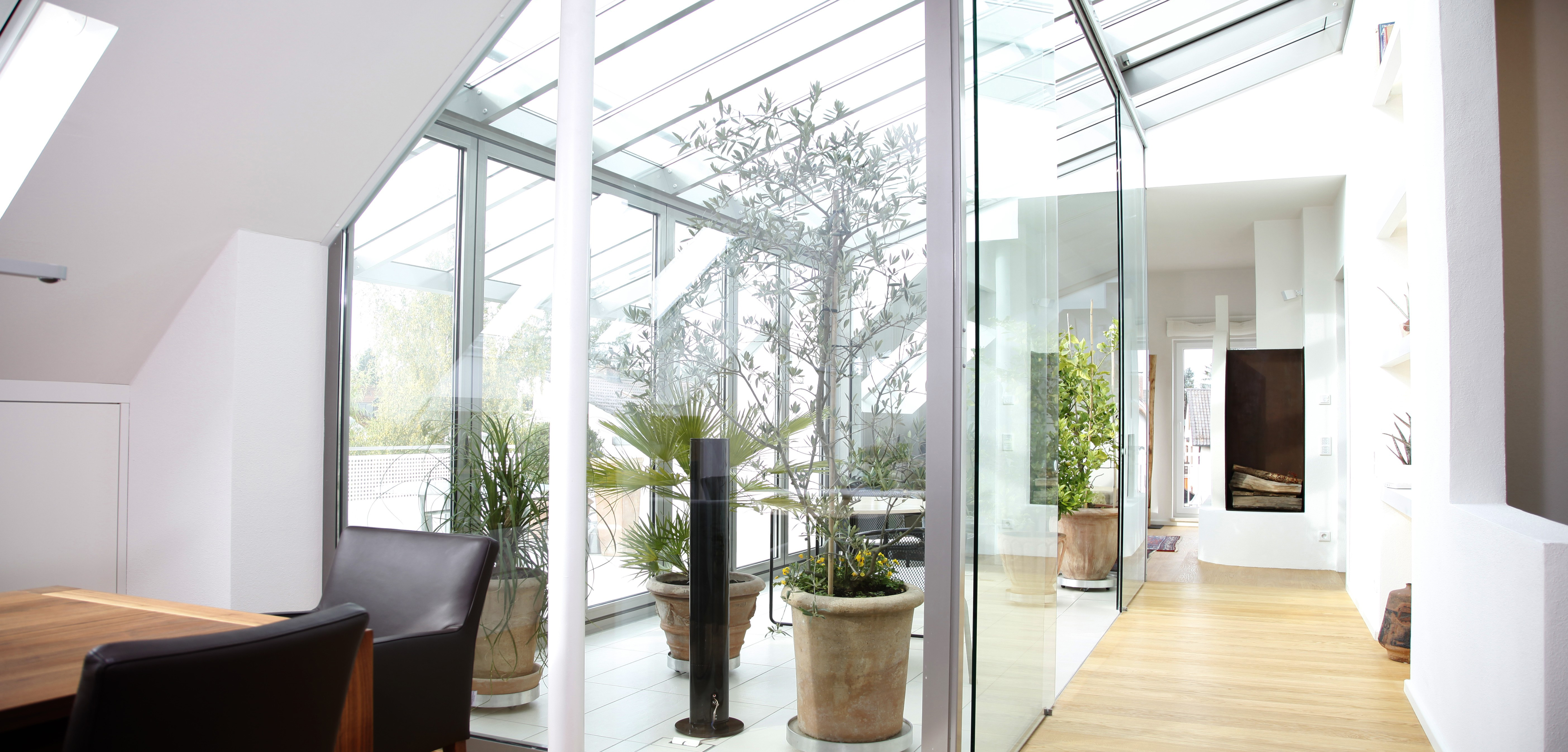 Ein moderner, lichtdurchfluteter Wohnbereich mit direkter Anbindung an einen hellen Wintergarten. Die raumhohen Glasflächen – inklusive Dachfenster und bodentiefer Verglasung – lassen reichlich Tageslicht herein. Auf einem hellen Holzboden stehen große, dekorativ bepflanzte Ton- oder Terrakottatöpfe, die dem Raum eine natürliche, frische Note verleihen. Links im Bild befindet sich ein eleganter Esstisch aus Holz, flankiert von dunklen, komfortablen Esszimmerstühlen. Im Hintergrund schimmert ein offener Flur, der in weitere Wohnbereiche führt. Die Gestaltung wirkt klar, luftig und harmonisch – mit fließenden Übergängen zwischen Innenraum, Essbereich und Wintergarten.