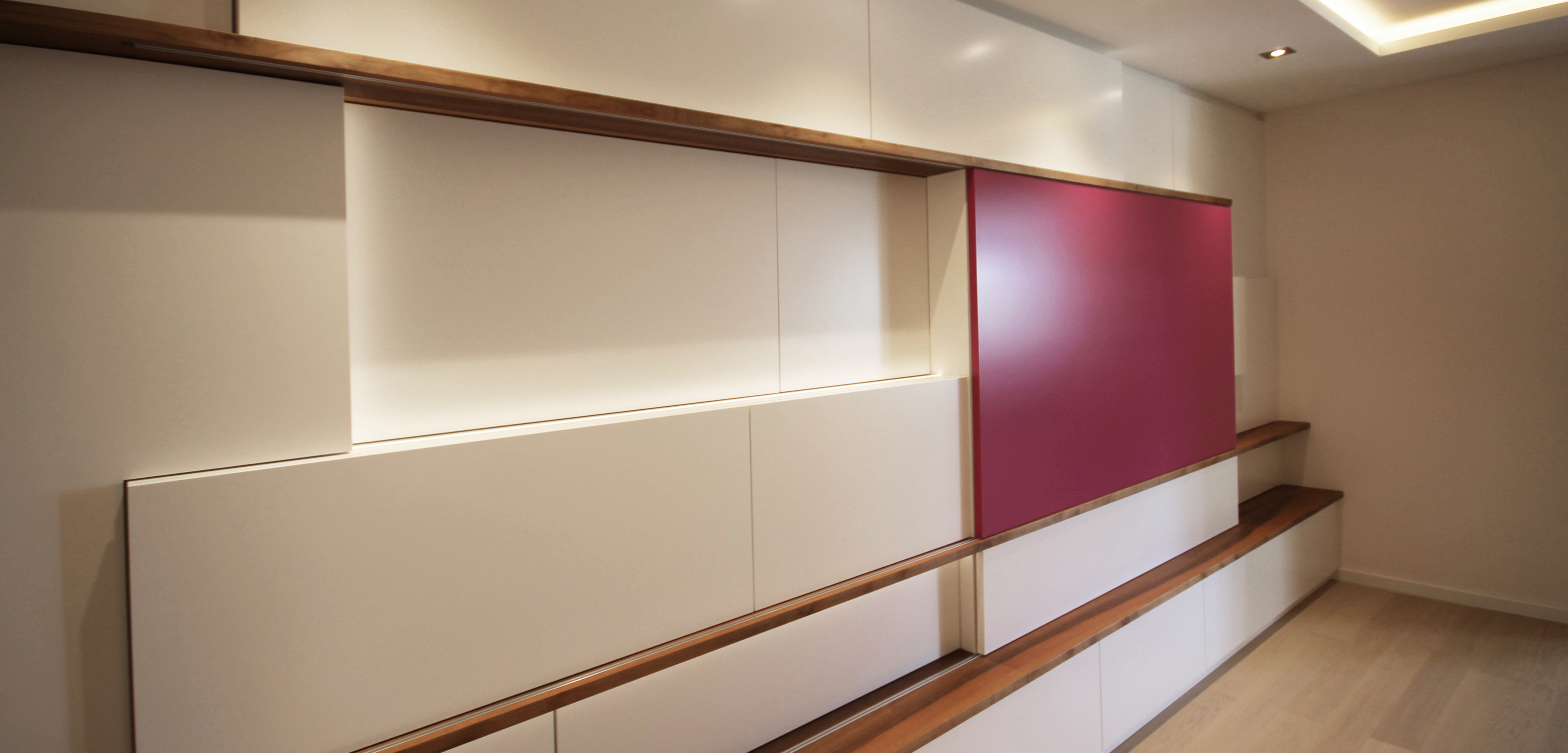 Maßgefertigter Wohnzimmerschrank mit verschiebbarer magentafarbener Front, hinter der ein Fernseher verborgen ist, kombiniert mit weißen Flächen und Holzleisten.