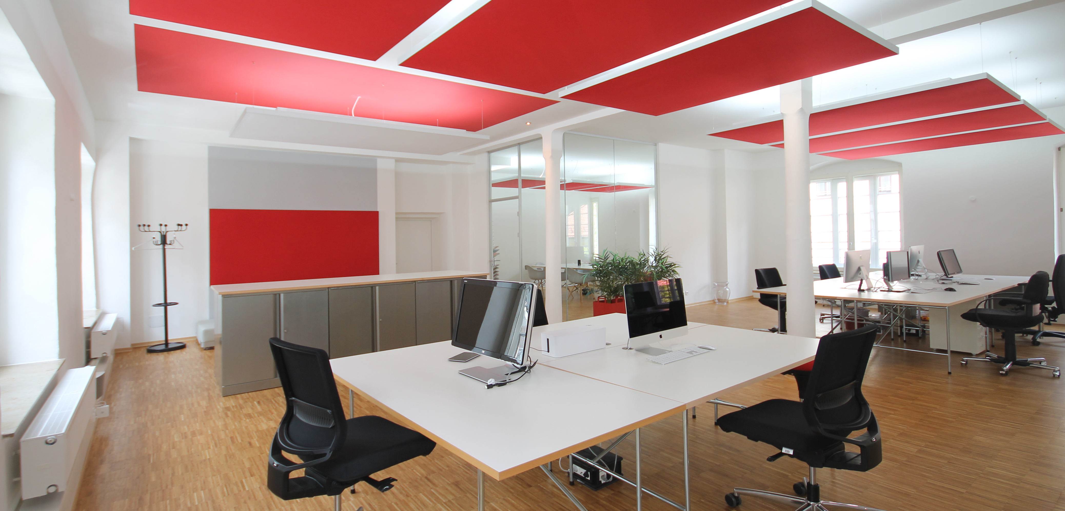 Offenes, helles Büro mit von eswerderaum geplanter Open-Space-Arbeitslandschaft: große weiße Schreibtische in Gruppen, ergonomische Drehstühle, Holzfußboden, rote Akustiksegel an der Decke und viel Tageslicht – ein Beispiel für moderne, funktionale Büroplanung von eswerderaum.