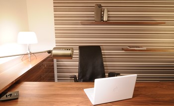 Der Arbeitsbereich mit Schreibtisch, Sideboard, Container aus Nussbaum wird durch eine Tapete mit Streifen betont. Bürostuhl aus Leder, Wandboard, Tischleuchte.
