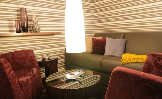 Die Lounge mit Ledersofa, Sessel, Couchtisch, Teppich, Stehleuchte wird von einer Tapete eingefasst. Edle Oberflächen und Materialien aus Nussbaum, Glas, Stoff.