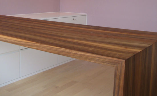 Der Schreibtisch liegt mit der Tischplatte auf dem Sideboard auf. Die Oberfläche besteht aus Nussbaum Furnier. Der Hängeschrank ist mit Einbauleuchten versehen.