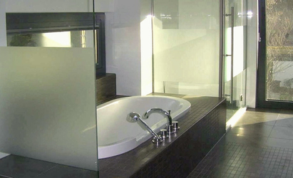 Mattierte Glasscheiben trennen Badewanne von WC-Sitz und Dusche. Die Wanne Oberfläche aus Emaille wird mit Standarmaturen aus Chrom auf dem Wannenrand bedient.