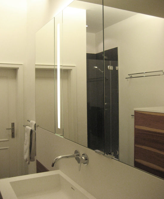 Der Spiegelschrank steht flächenbündig auf der Rückwand des Waschtisches auf. Hinter den verspiegelten Glastüren finden sich Gefache für Kosmetika, integrierte Spiegelbeleuchtung.