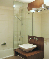Der freihängende Waschtisch mit Aufsatzbecken, Wandarmatur und Spiegelschrank schließt an die Duschabtrennung aus Glas an. Diese steht auf dem Rand der Badewanne auf.