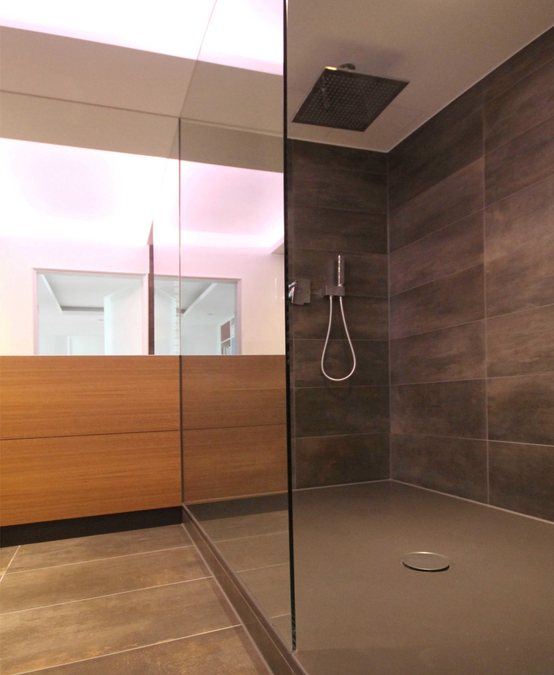 Die raumhohe Duschtrennwand aus Glas steht auf dem Rand der Duschwanne auf. Die Regenbrause ist an der Raumdecke installiert. Die Wandfliesen bestehen aus Feinsteinzeug.