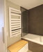 Ein Falttür aus Glas ermöglicht die Nutzung der Badewanne als Dusche, aufgeklappt parkt sie seitlich vor der Fliesenwand, eine Sitzbank dient als Sammelbehälter für Schmutzwäsche.
