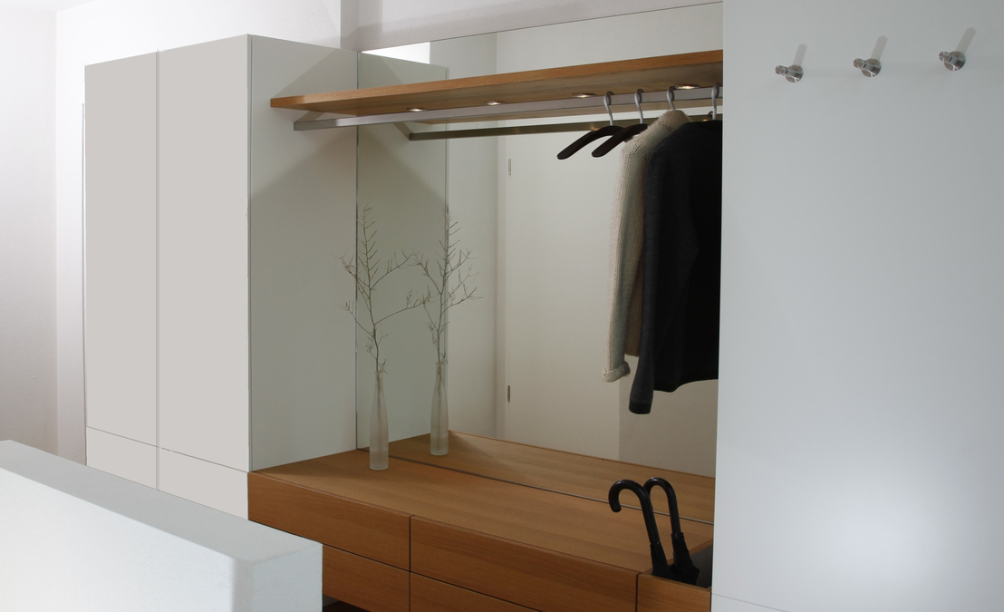 Ein Schuhschrank mit lackierten Oberflächen ist Pendant zur Wandverkleidung mit demselben Material, die Kleiderhaken trägt. Dazwischen Spiegel und Hängeboard. 