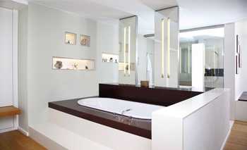 Badewanne und Waschtisch sind durch eine halbhohe Mauer vom Raum getrennt. In die Wand eingelassene Glaskästen sind beleuchtet. Spiegel sind Maßanfertigung.