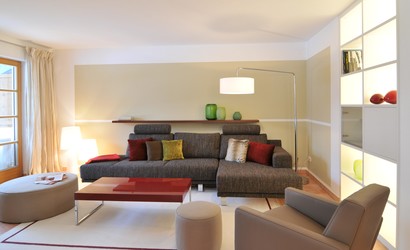 Das Wohnzimmer ist mit Ecksofa, Sessel, Hocker Couchtisch, Stehleuchte und einem Teppich ausgestattet. Das Regal ist passgenau eingebaut und beleuchtet.