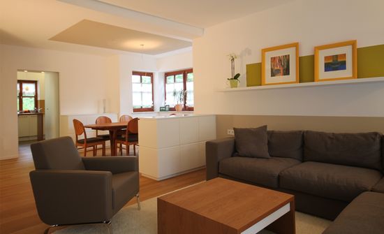 Der Wohnraum mit Ecksofa, Couchtisch, Sessel, Teppich und Wandboard wird durch ein Sideboard vom Essbereich getrennt. Der Dielenboden läuft bis in die Küche.