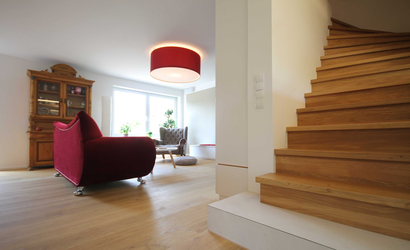 Der Fußboden ist mit einem Parkett aus Landhausdielen ausgeführt, das Antrittspodest der Treppe ist mit Fliesen belegt, die Schirmleuchte im Wohnzimmer passt zum Sofa. 