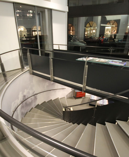 Eine automatische Glasschiebetür bildet den Haupteingang des Ladens. Ein Steg führt über die Treppenöffnung in den Verkaufsraum. Der graue Boden besteht aus Kunstharz.