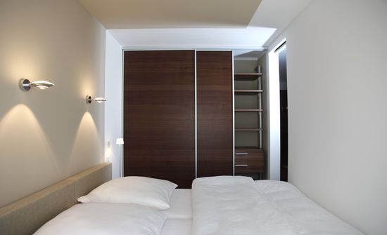 Betthaupt und Fußende des Doppelbetts passen zwischen die Raumwände. Zugang zu beiden Seiten über Schiebetüren, Wandfarbe und Wandleuchten, Kleiderschrank.