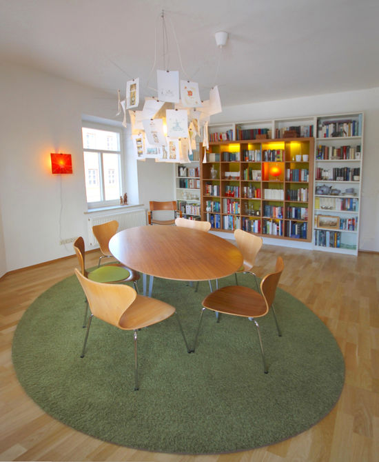 Der ovale Tisch mit sechs Stühlen, der ovale Teppich sowie die Pendelleuchte markieren das Zentrum des außerrechtwinkligen Raumes. Die Bücher-Regalwand ist hinterleuchtet.