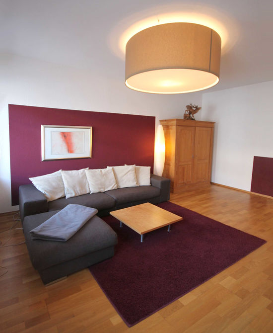 Eine große Deckenleuchte mit Stoffbespannung und Diffusor wirkt wie eine Lichtdecke und verdichtet die Raumzone mit dem Sofa, dadurch wird Behaglichkeit und Atmosphäre erzeugt.