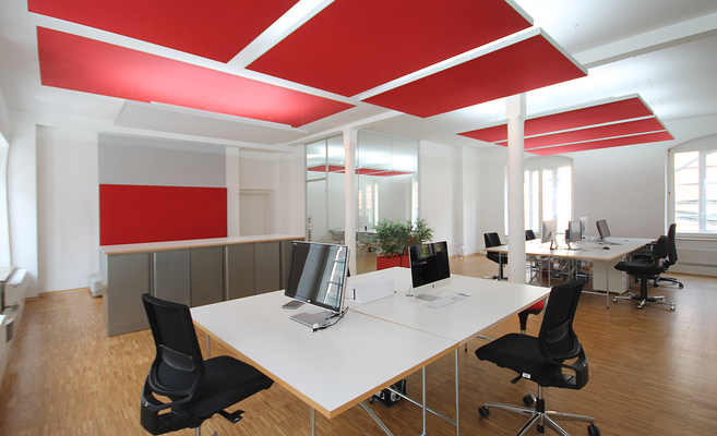 Offenes, helles Büro mit von eswerderaum geplanter Open-Space-Arbeitslandschaft: große weiße Schreibtische in Gruppen, ergonomische Drehstühle, Holzfußboden, rote Akustiksegel an der Decke und viel Tageslicht – ein Beispiel für moderne, funktionale Büroplanung von eswerderaum.