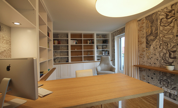 Innenansicht eines stilvollen Home Offices mit einer großzügigen Bibliothek. An den Wänden erstrecken sich hohe Bücherregale, die mit einer Vielzahl von Büchern gefüllt sind und eine entspannte, inspirierende Arbeitsatmosphäre schaffen. In der Mitte des Raumes befindet sich ein moderner Schreibtisch mit einem ergonomischen Stuhl, der vor einem Fenster steht, das viel natürliches Licht hereinlässt. Der Raum ist ideal für produktives Arbeiten im Home Office, mit einem ausgewogenen Mix aus Funktionalität und gemütlichem Design.