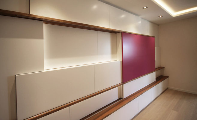 Maßgefertigter Wohnzimmerschrank mit verschiebbarer magentafarbener Front, hinter der ein Fernseher verborgen ist, kombiniert mit weißen Flächen und Holzleisten.